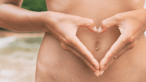 A fit woman's toned midsection, with her hands gently forming a heart shape around her belly button, symbolizing love and care for gut health. This image is a testament to the importance of maintaining a healthy digestive system for overall wellness.
