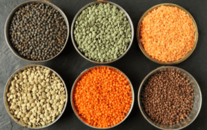 Six types of lentils arranged side by side for comparison. From left to right, we see the Green Lentil, commonly used in European dishes, Puy Lentils, a French variety with a peppery taste, Red Lentils, which are favored for their quick cooking time and are often seen in Indian dals, Yellow Lentils, similar to red in taste and texture, Black Beluga Lentils, known for their firm texture, and Brown Lentils, the most versatile variety used in a range of cuisines.