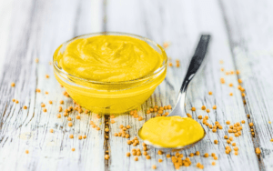 The image showcases a glass bowl filled with piquant mustard, accompanied by a spoon and it's scattered seeds, all presented on a rustic table.